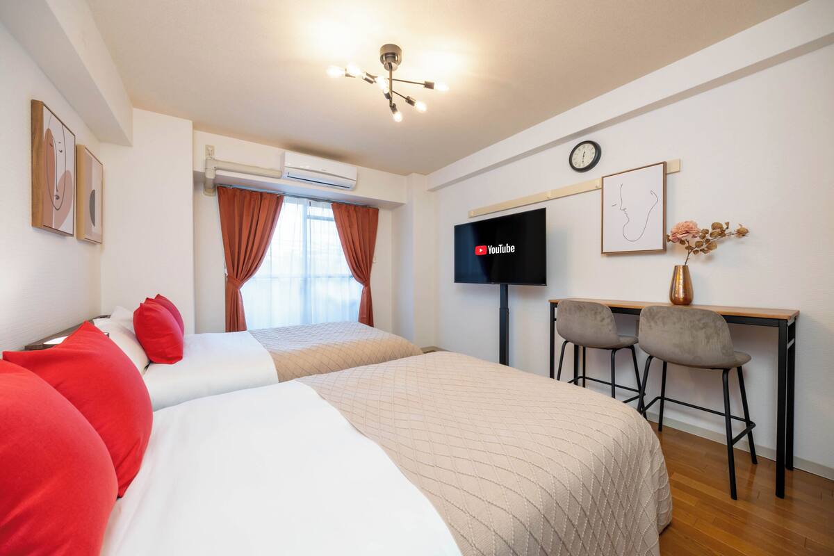 A hotel room with a bed, a desk with two chairs, a flat-screen TV, an air conditioning unit, and a clock on the wall.