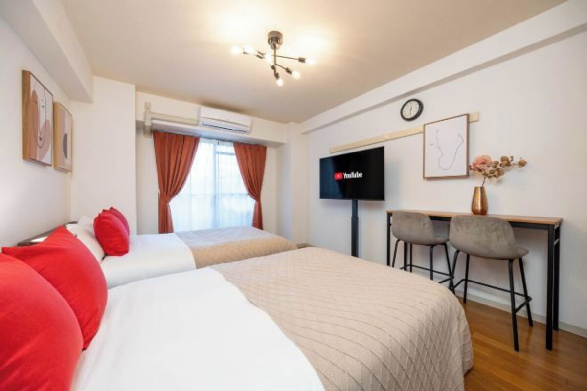 A hotel room with a bed, a desk with two chairs, a TV, and a clock on the wall.