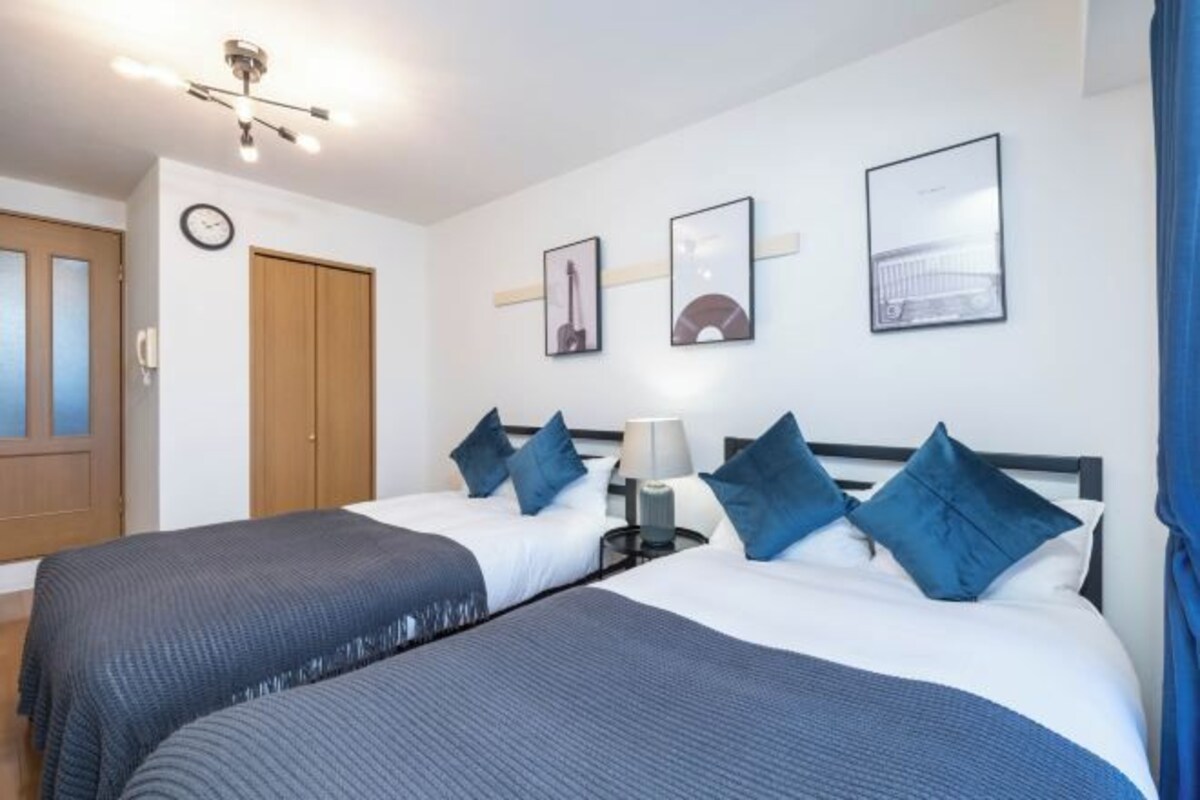A hotel room with two beds, a wooden door, a clock, and framed pictures on the wall.