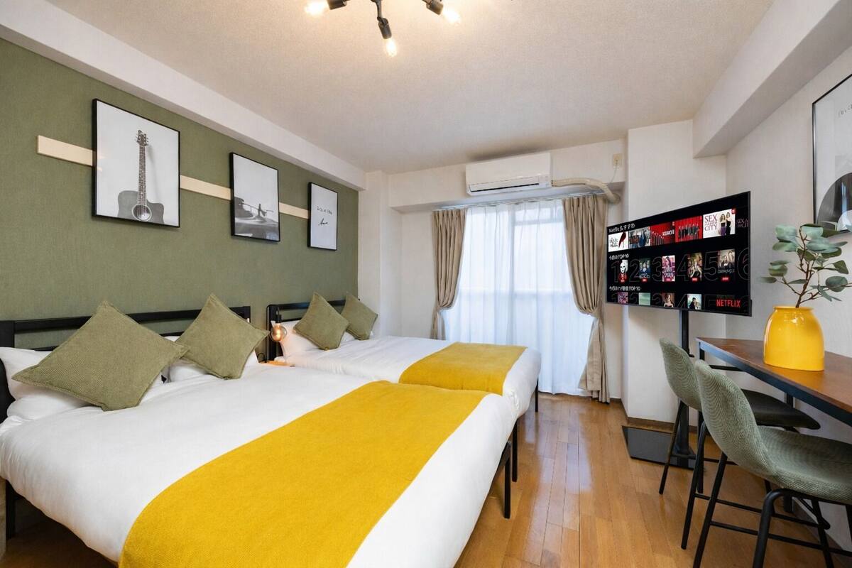 A hotel room with two beds, a desk, a chair, an air conditioning unit, and a wall-mounted TV displaying movie options.
