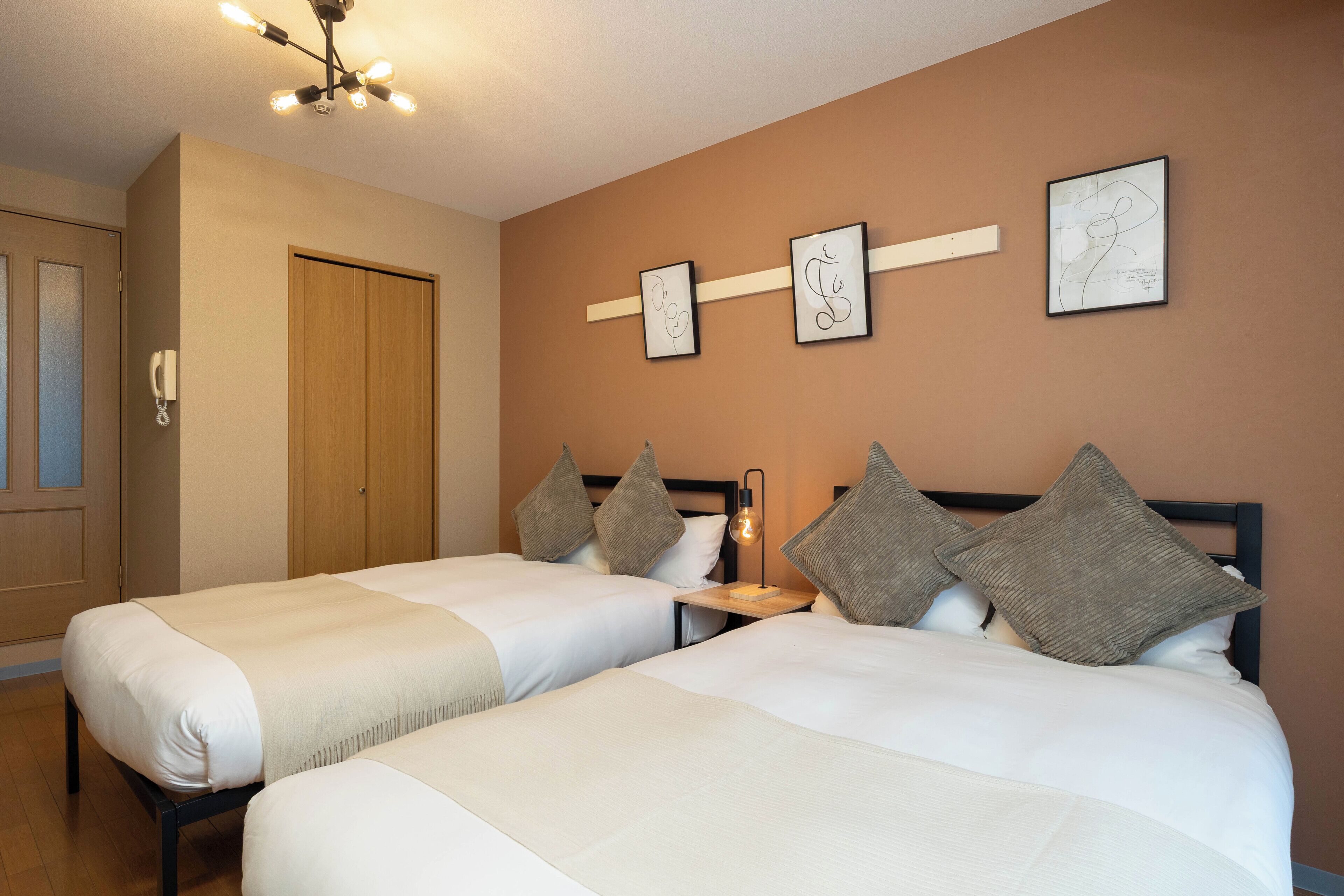 A hotel room with two beds, a wooden wardrobe, a bedside table with a lamp, and artwork on the wall.
