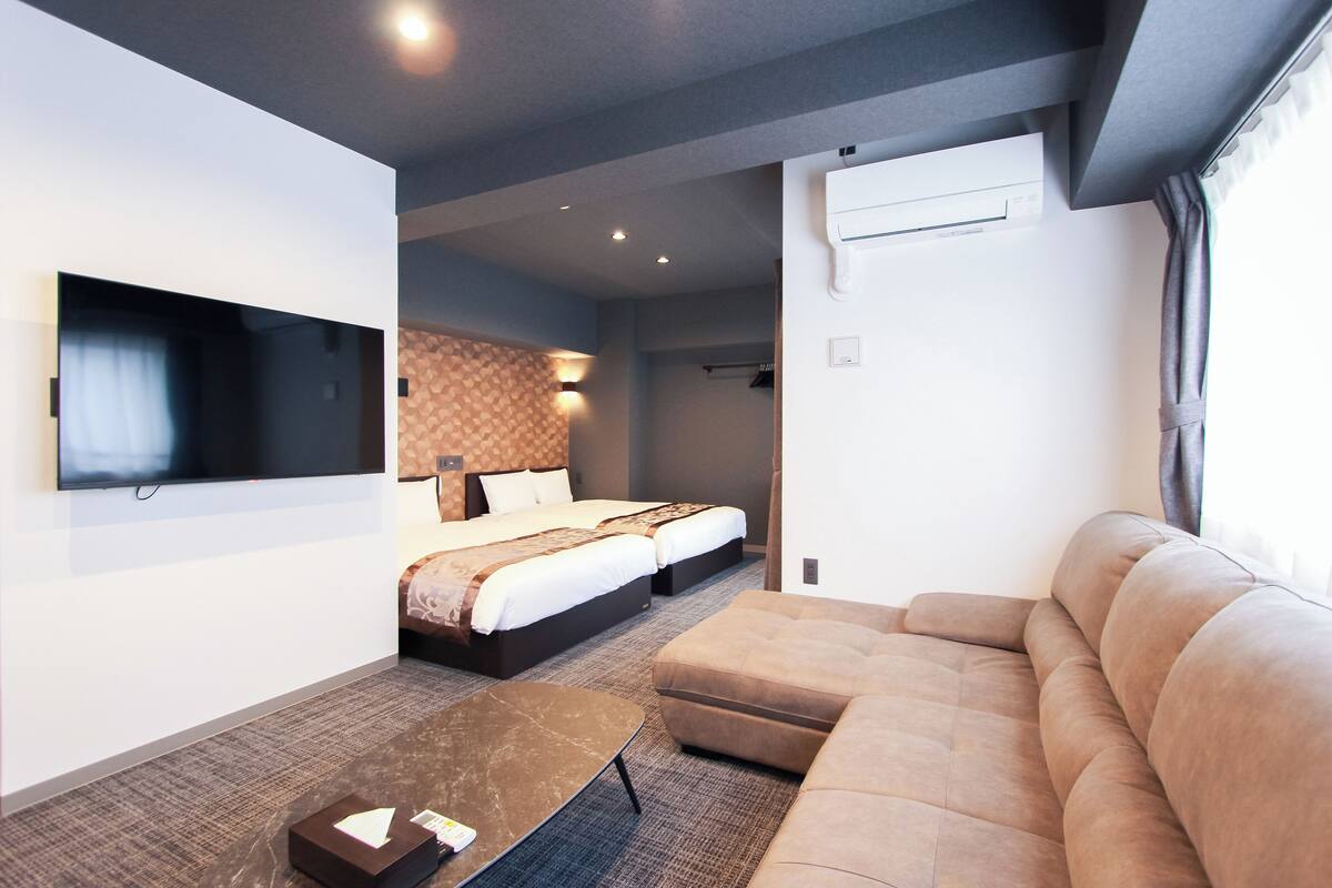 A modern hotel room with a flat-screen TV mounted on the wall, a sofa, a bed with two pillows, and a small table.
