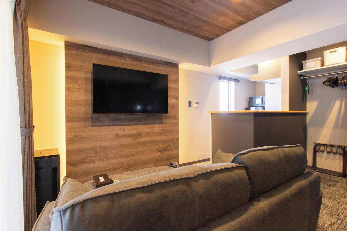 A modern living room with a large flat-screen TV mounted on a wooden wall, a sofa, and a kitchen area visible through an open doorway.