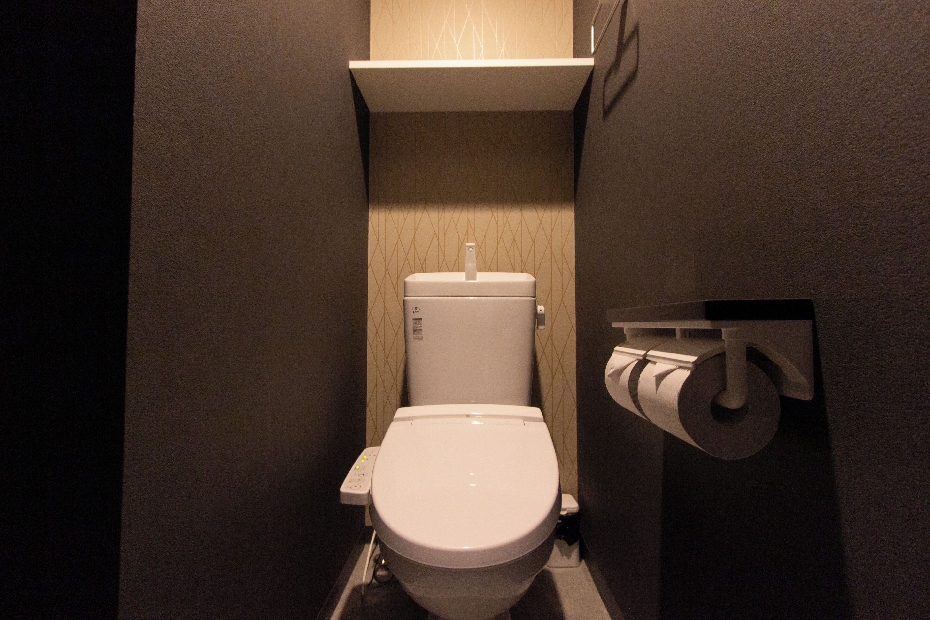 A small, compact bathroom with a toilet, a toilet paper holder, and a wall-mounted shelf.