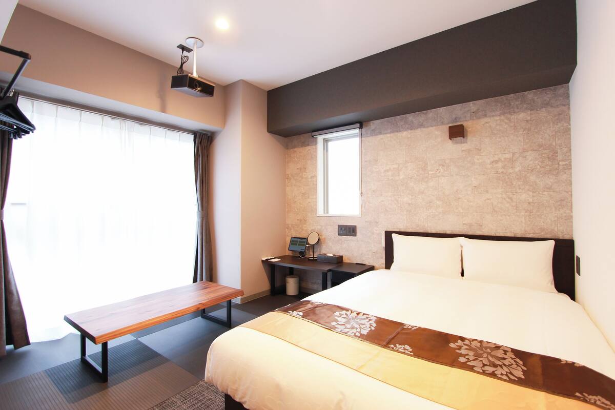A hotel room with a large bed, a desk with a computer, a bench, and a window with curtains.
