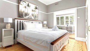 A bedroom with a large bed, two bedside tables, a painting, and a view of the outdoors. - Luxury Villas Shelley St (Pet Friendly) (Elwood)