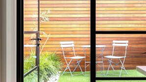 A sliding glass door leading to an outdoor patio with a wooden wall, two white folding chairs, and a small table. - Luxury Villas Shelley St (Pet Friendly) (Elwood)