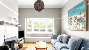 A living room with a fireplace, a sofa, a coffee table, and a large window with blinds. - Luxury Villas Shelley St (Pet Friendly) (Elwood)