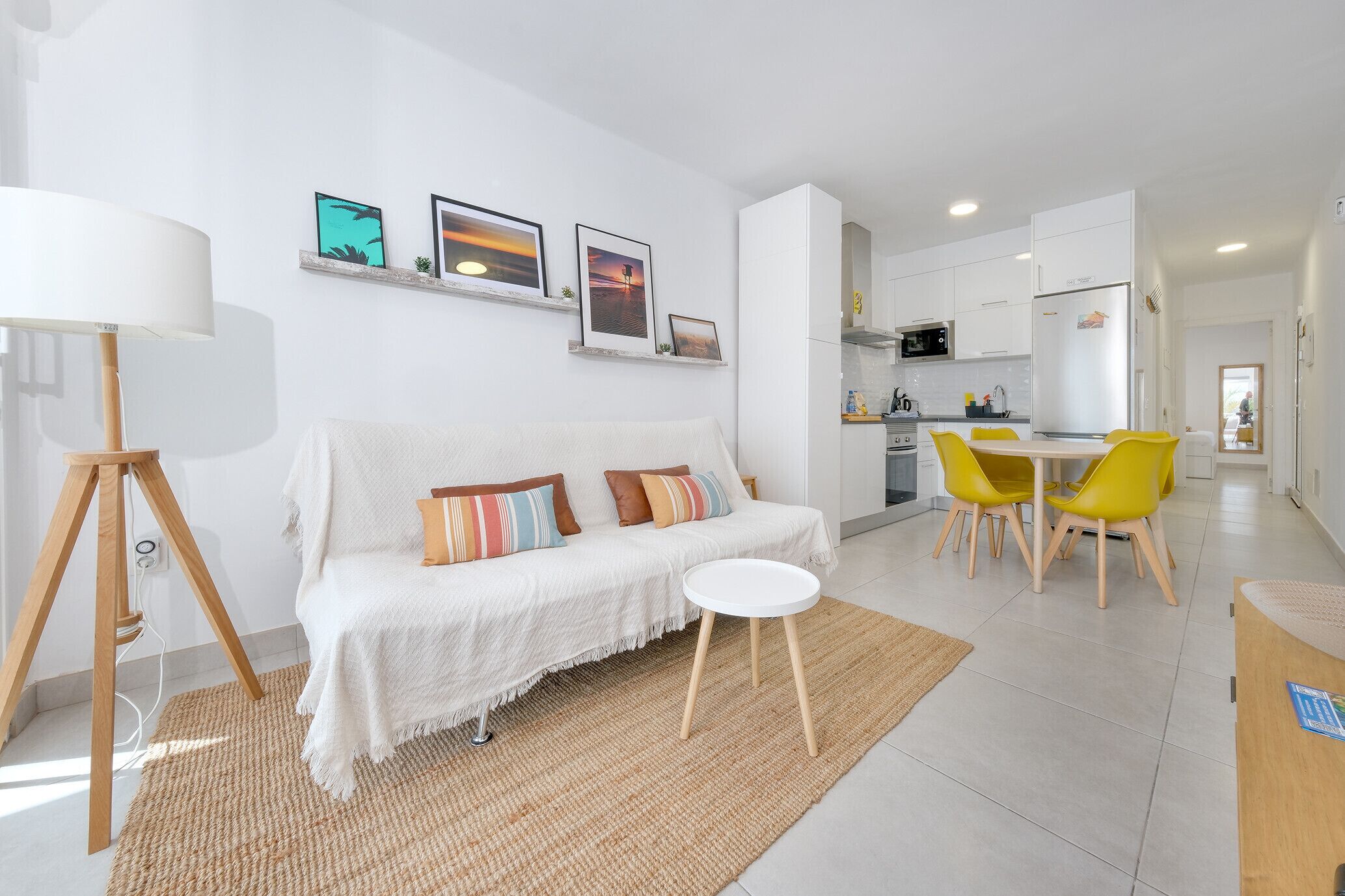 Uma sala de estar moderna com um sofá branco, uma mesinha redonda pequena e um espaço de refeições com cadeiras amarelas. É possível ver uma cozinha ao fundo, com armários brancos e um micro-ondas.