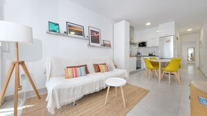 Un salon moderne avec un canapé blanc, une petite table ronde et un coin repas avec des chaises jaunes. On aperçoit au fond une cuisine avec des meubles blancs et un four à micro-ondes.