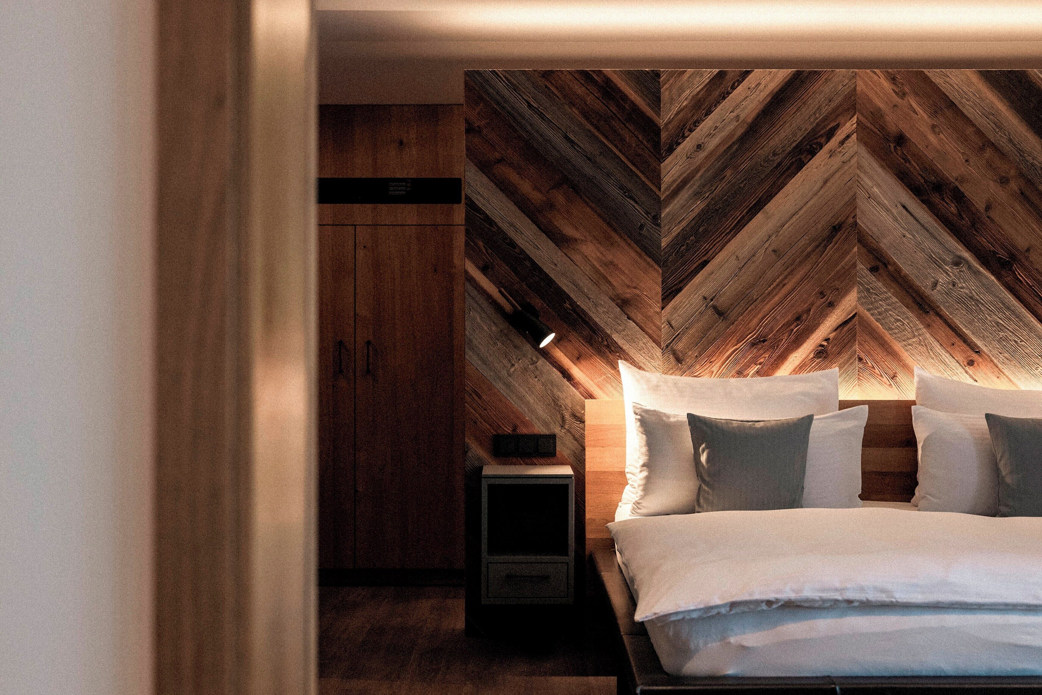Un dormitorio con techo de madera con patrón de espiga, cama con ropa de cama blanca y mesita de noche con lámpara.