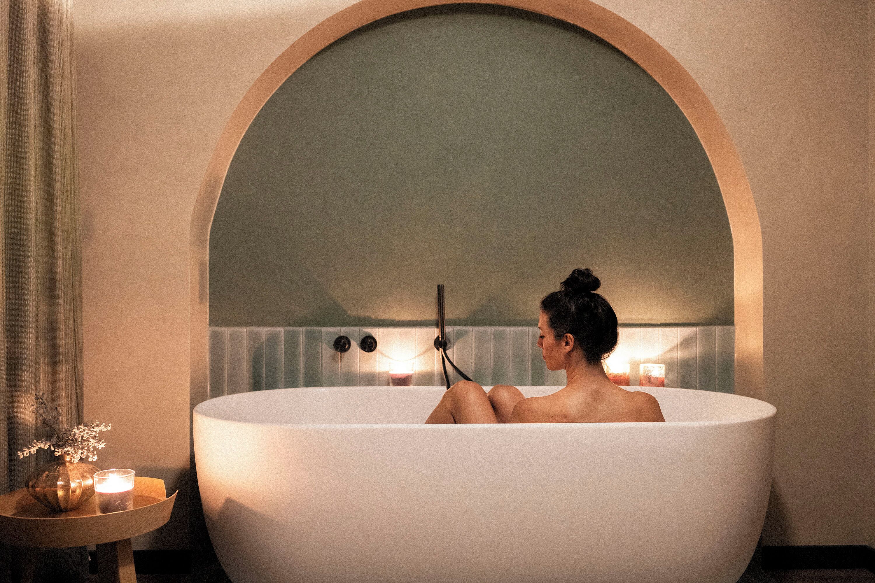 A person relaxing in a large, white bathtub with a wooden side table beside it, holding a book.