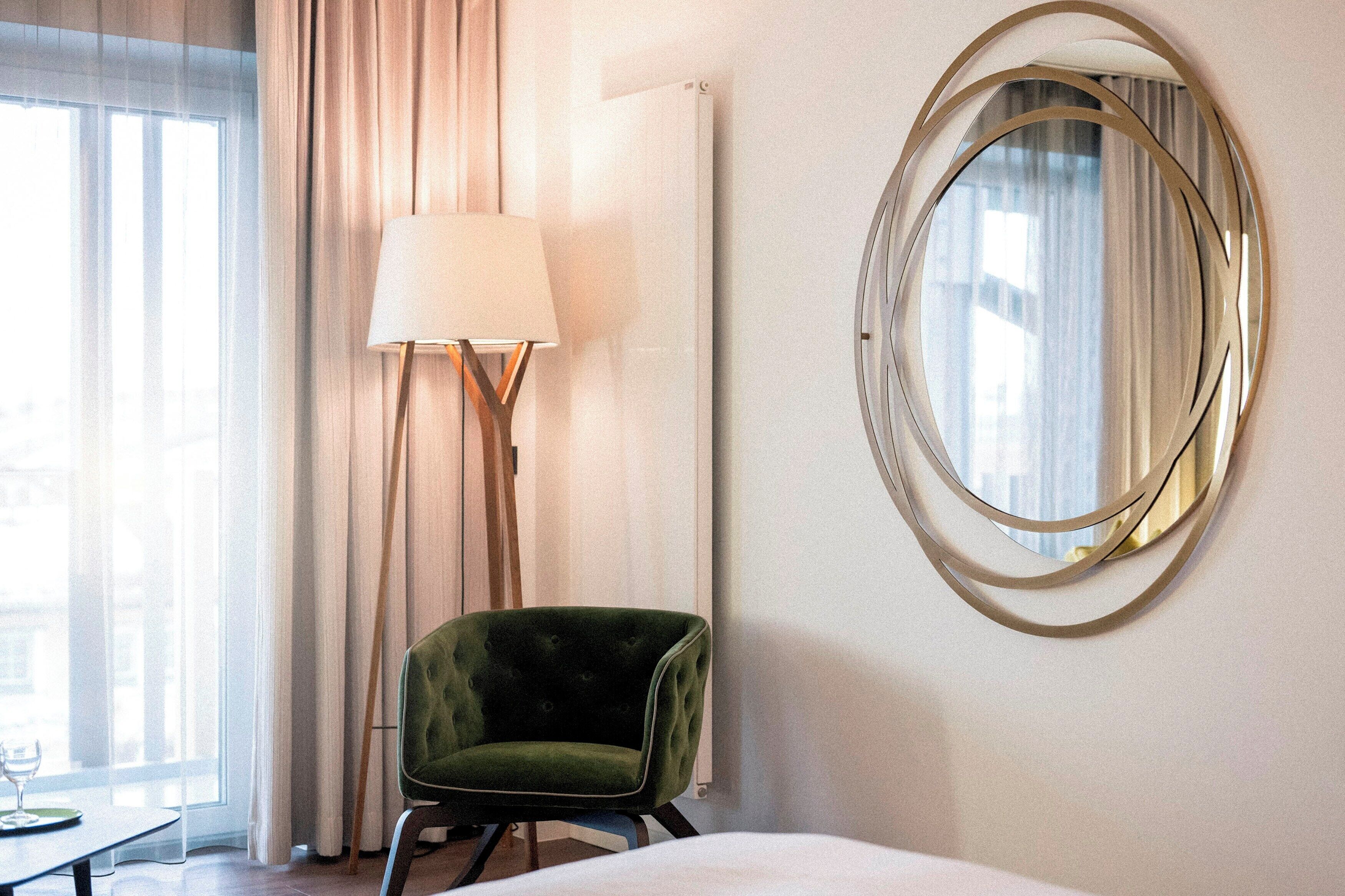 A hotel room with a green armchair, a floor lamp, a round mirror, and a bed.