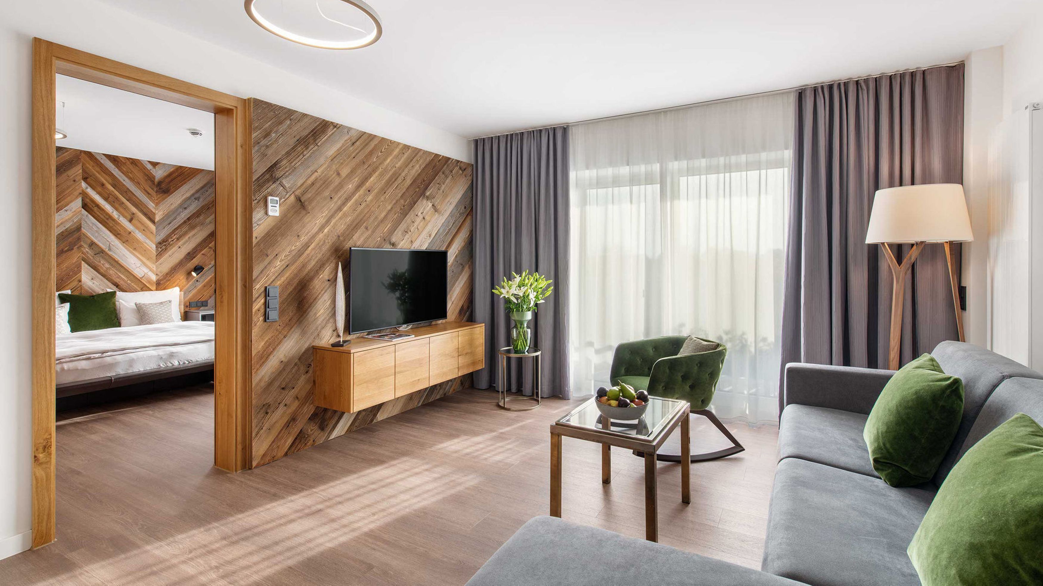 Een moderne hotelkamer met een houten visgraatwand, een flatscreen televisie, een groene fauteuil, een grijze bank en een nachtkastje met een bloemenvaas.