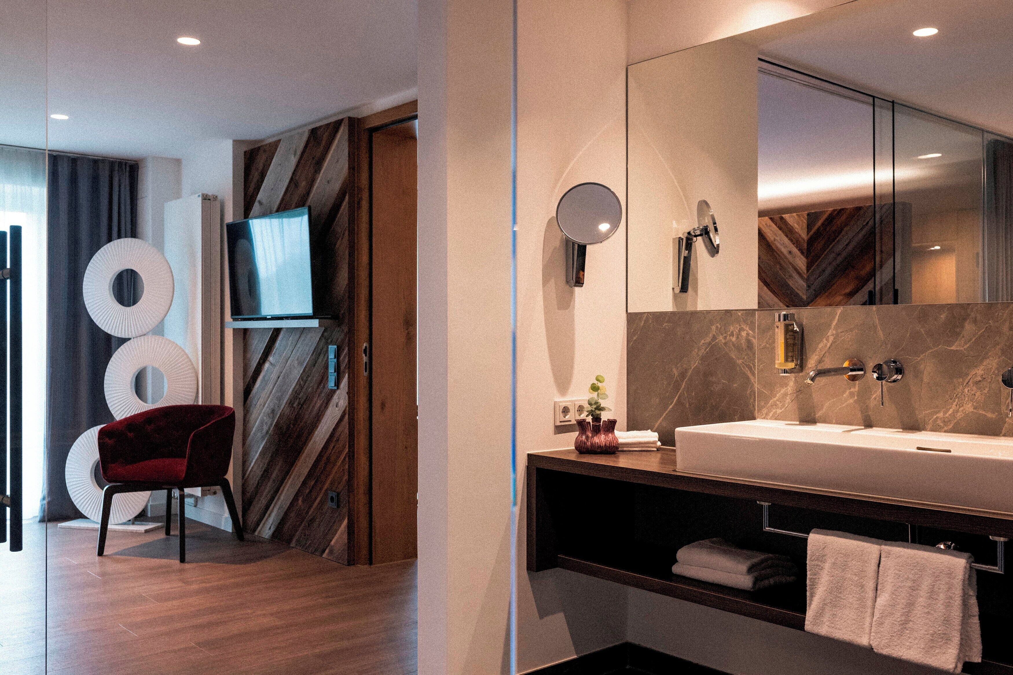 Un baño moderno con un espejo grande, un lavabo y una ducha. En la habitación contigua hay una silla roja y un televisor.