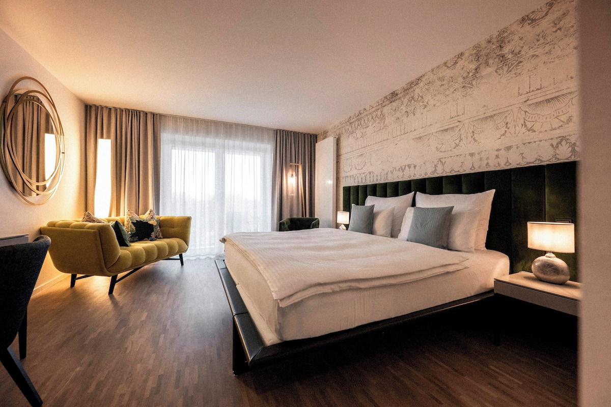 A hotel room with a large bed, a sofa, a chair, and a nightstand with a lamp.