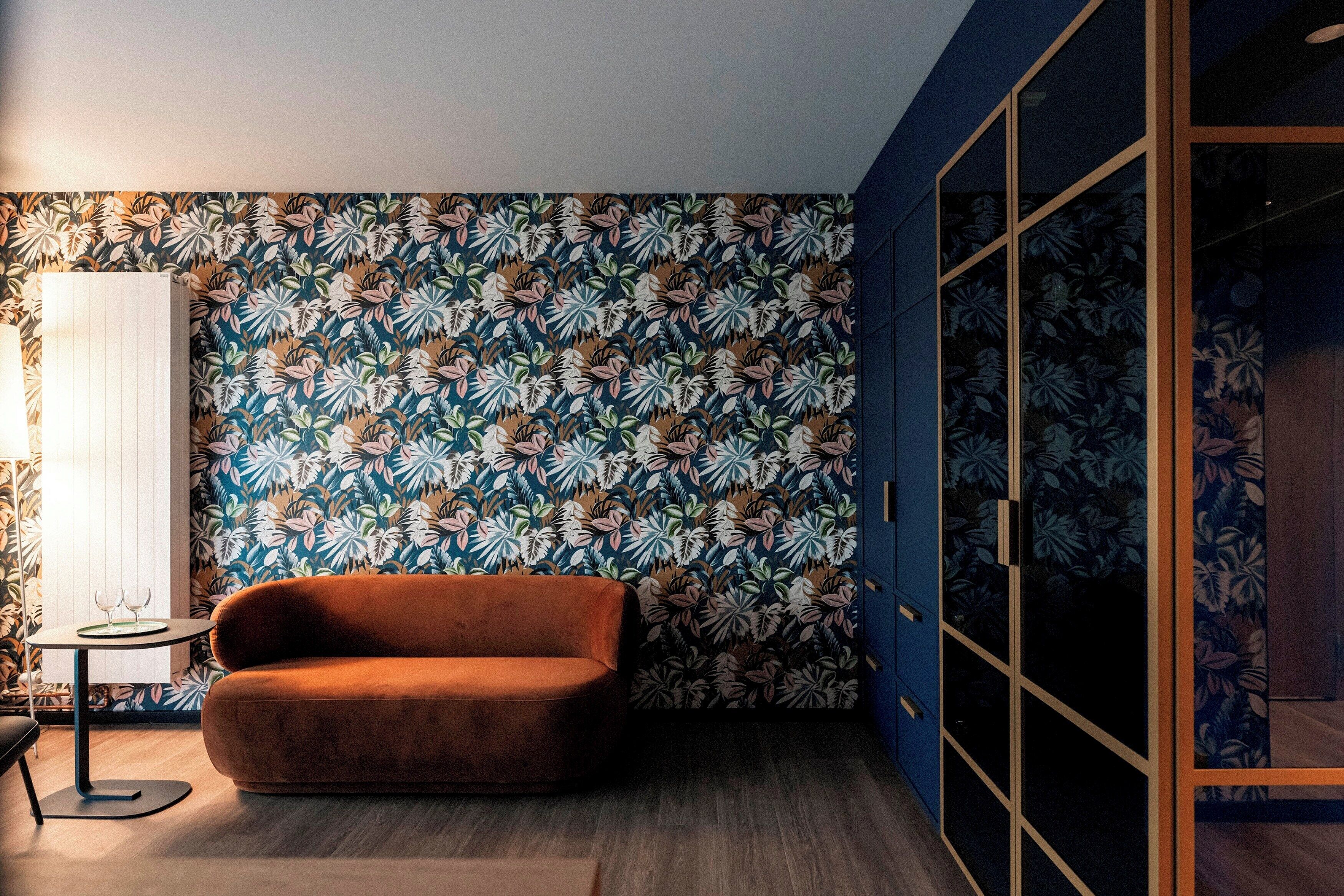 A room with a patterned wallpaper, a brown sofa, a small table with a lamp, and a wardrobe with a glass door.