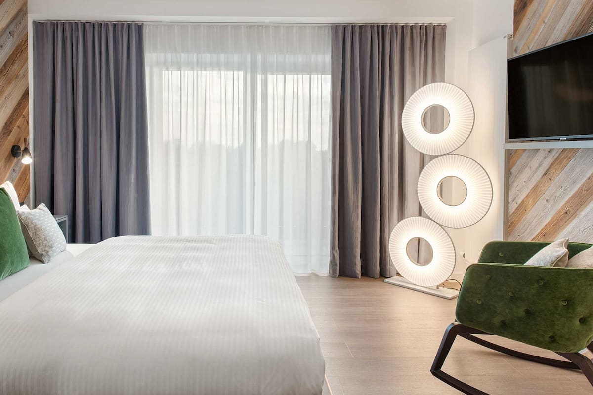 A modern hotel room with a large bed, grey curtains, a green armchair, and a wall-mounted TV.