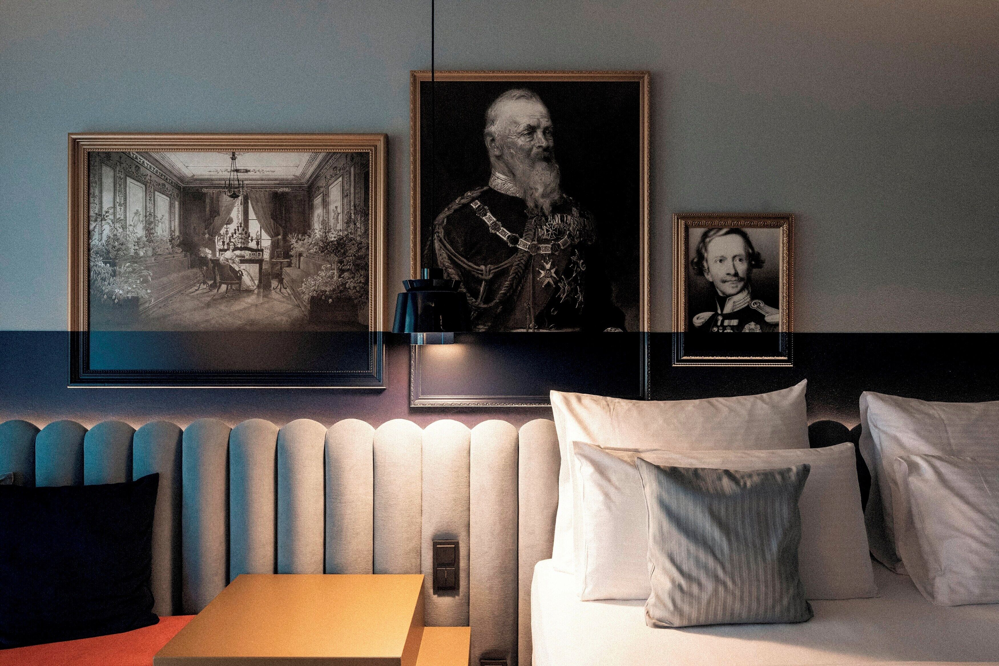 Ein Hotelzimmer mit Bett, Nachttisch und Heizkörper. An der Wand hängen gerahmte Bilder.