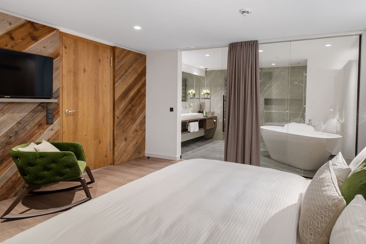 A modern hotel room with a large bed, a green armchair, a flat-screen TV, wooden paneling, a bathroom with a bathtub, and a shower.