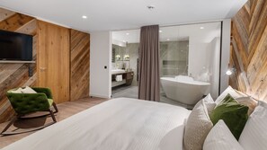 Moderni hotellihuone, jossa on suuri sänky, vihreä nojatuoli, litteänäyttöinen televisio, puupanelointi, kylpyammeella varustettu kylpyhuone ja suihku.