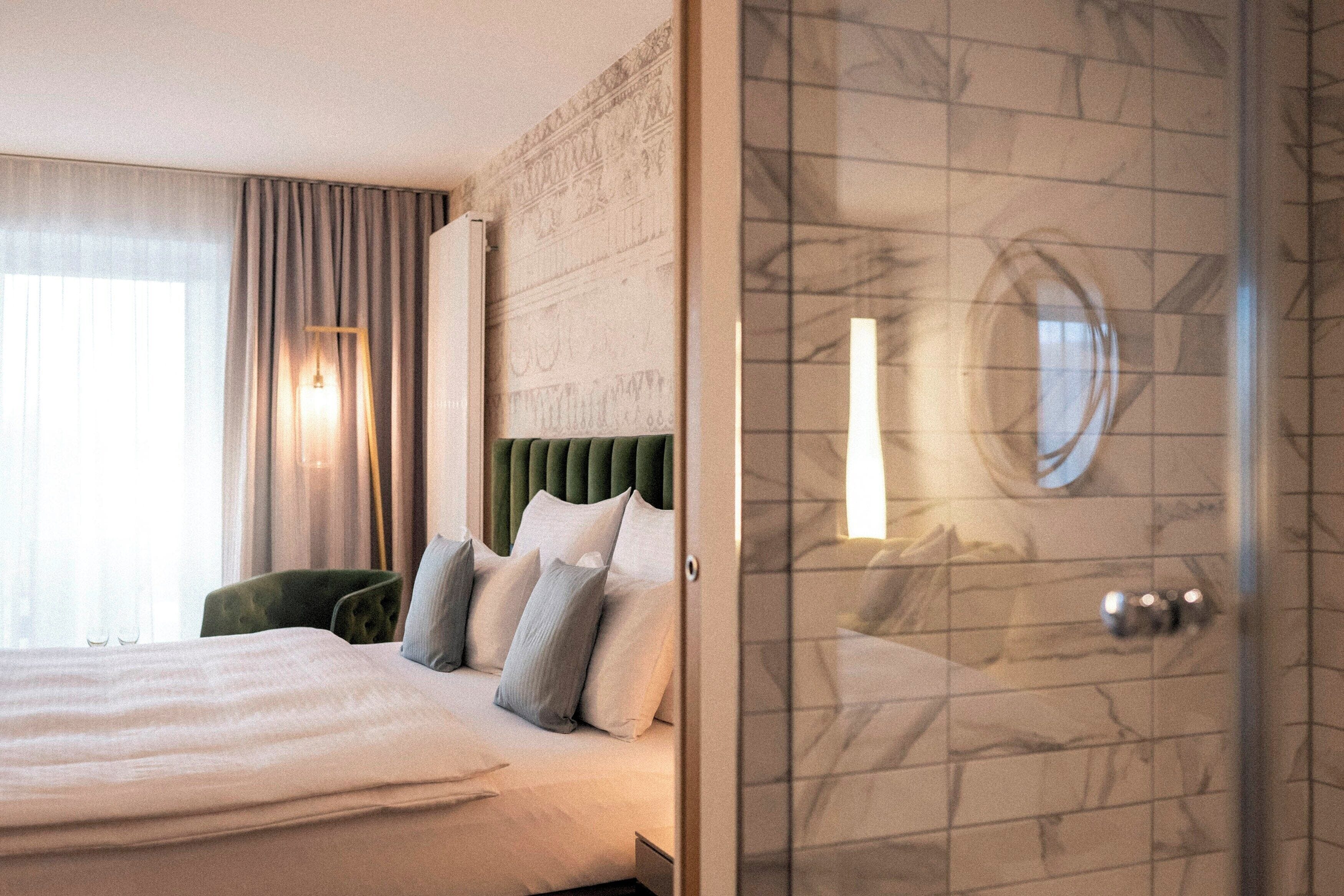 A hotel room with a bed, a green upholstered chair, a wall with a decorative pattern, and a tiled shower area.
