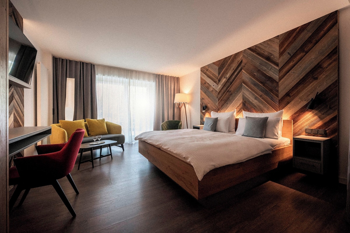 A modern hotel room with a large bed, a desk, a chair, and a sofa.