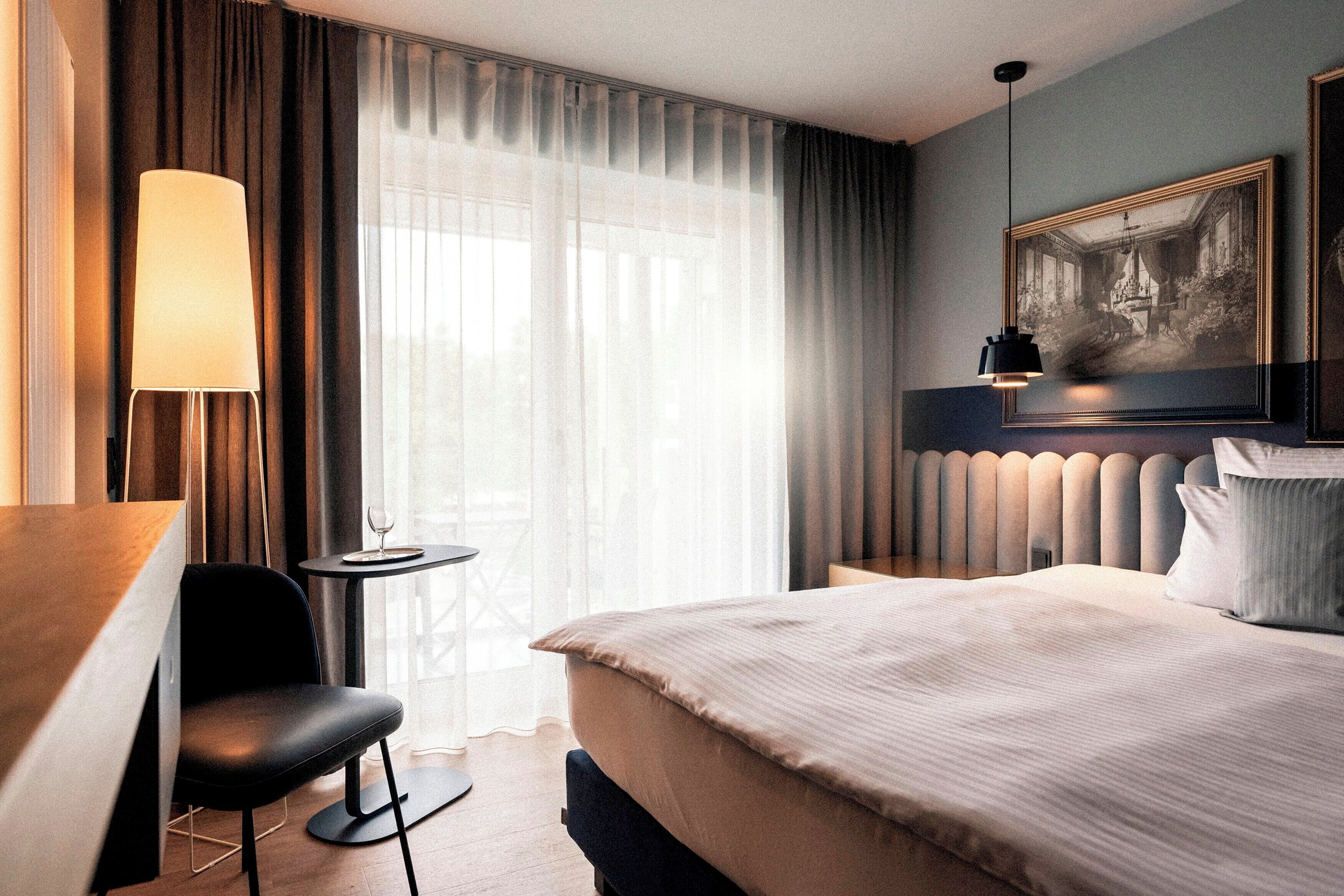 Ein Hotelzimmer mit einem Bett, einem Schreibtisch, einem Sessel, einem kleinen Tisch, einer Lampe, einem Fenster mit Vorhängen und einem gerahmten Bild an der Wand.