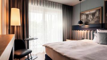 Ein Hotelzimmer mit einem Bett, einem Schreibtisch, einem Sessel, einem kleinen Tisch, einer Lampe, einem Fenster mit Vorhängen und einem gerahmten Bild an der Wand.