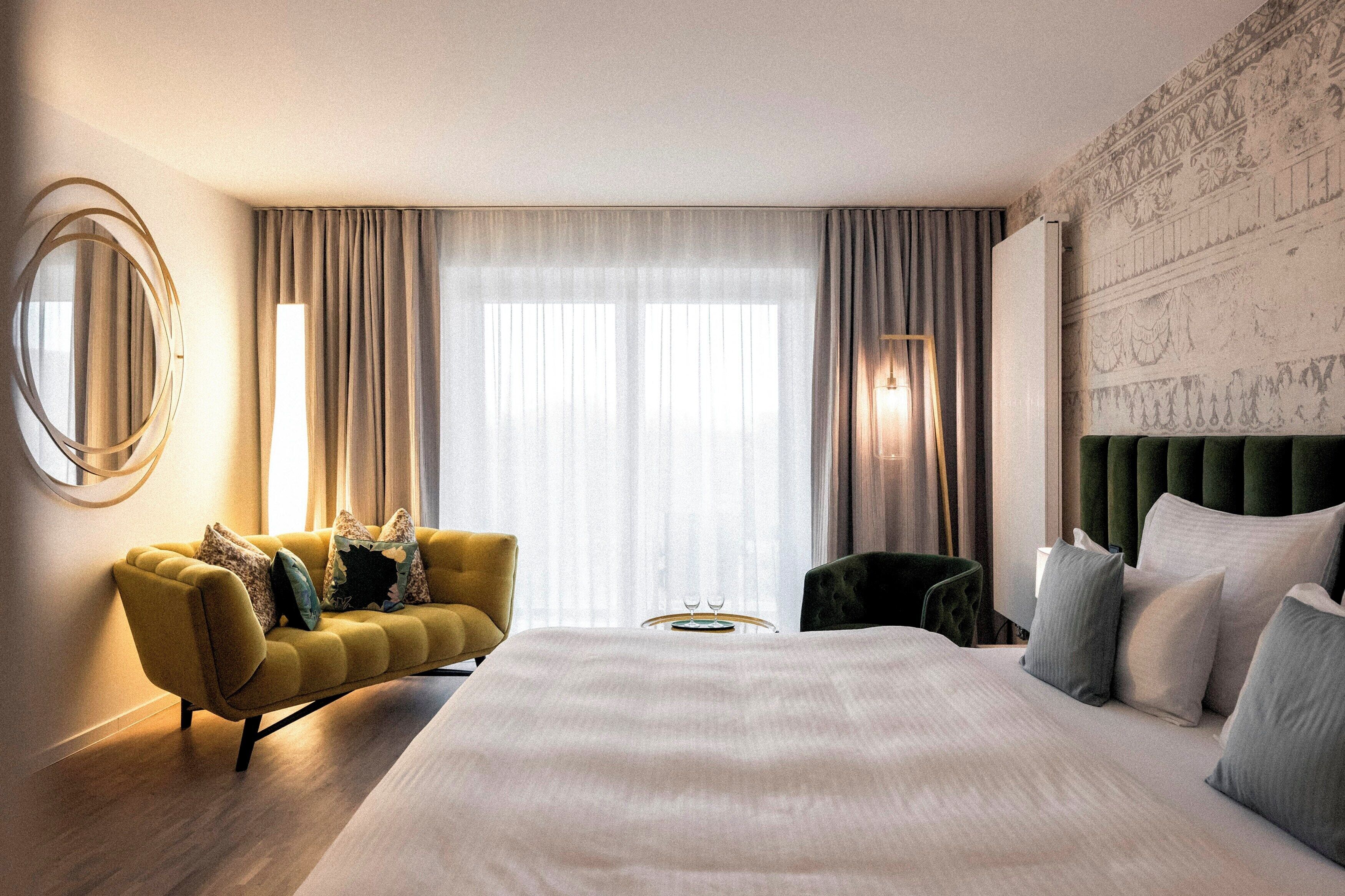 A hotel room with a large bed, a sofa, a chair, a round mirror, and a wall with a decorative pattern.