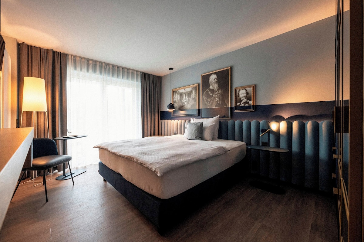 A modern hotel room with a large bed, a desk, and a chair. There are three framed pictures on the wall, a lamp, and a window with curtains.