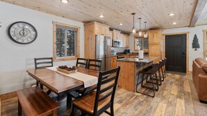 A modern kitchen with wooden cabinets, a central island, and a dining area with a wooden table and chairs. - The Black Bear Escaped! Hot Tub, Fire Pit , Yellowstone, Golf Course! (Island Park)