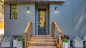 A modern building entrance with a wooden staircase, concrete planters with plants, and a grey door. - Mill Creek by AvantStay | Modern Design, Patio, Hot Tub (Healdsburg)