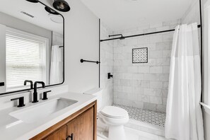 Ein modernes Badezimmer mit einem weißen Waschbecken, schwarzen Wasserhähnen, einem Holzschrank, einer weißen Toilette, einer Glasdusche und einem weißen Duschvorhang.