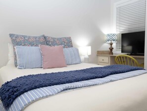 A neatly made bed with floral and striped pillows, a blue knitted throw, a bedside lamp, a television, and a chair.