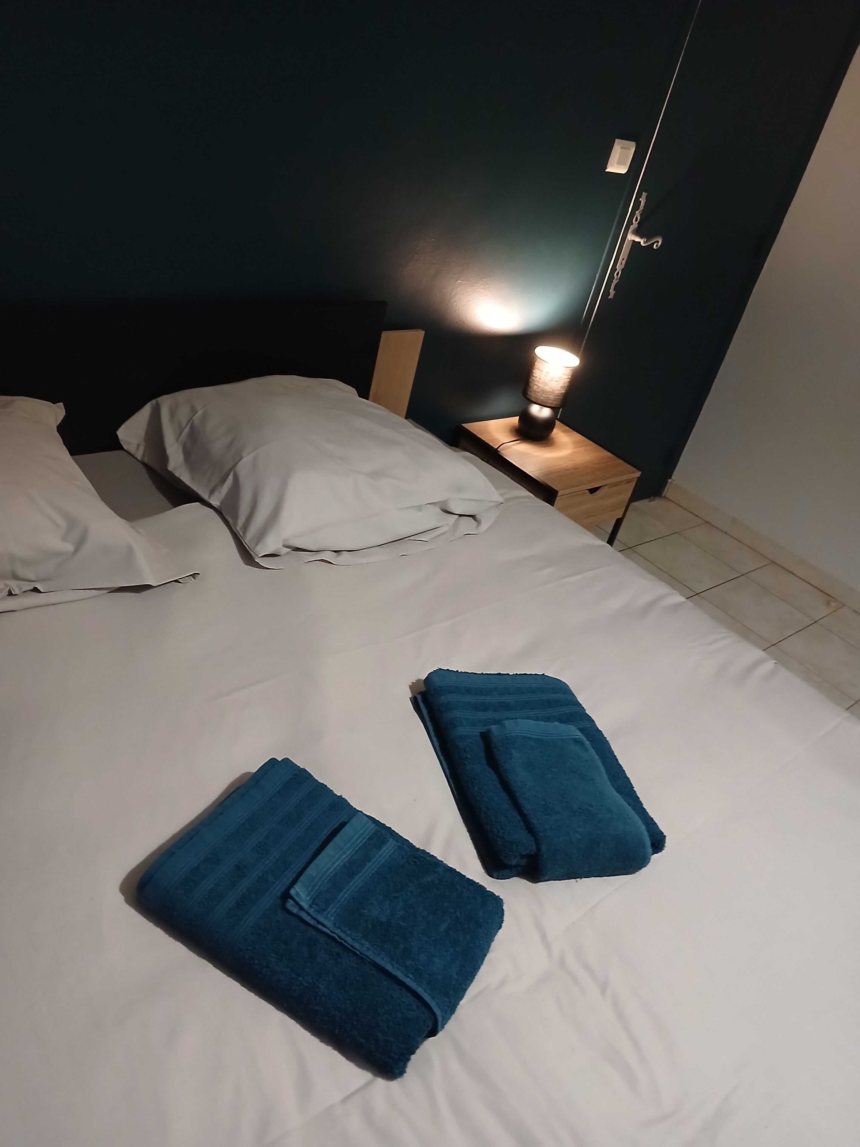 A neatly made bed with white sheets and two folded blue towels. There is a bedside table with a lamp and a wall-mounted light.