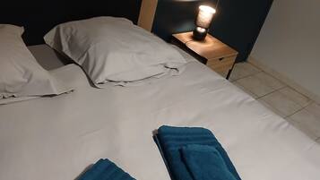 A neatly made bed with white sheets and two folded blue towels. There is a bedside table with a lamp and a wall-mounted light.