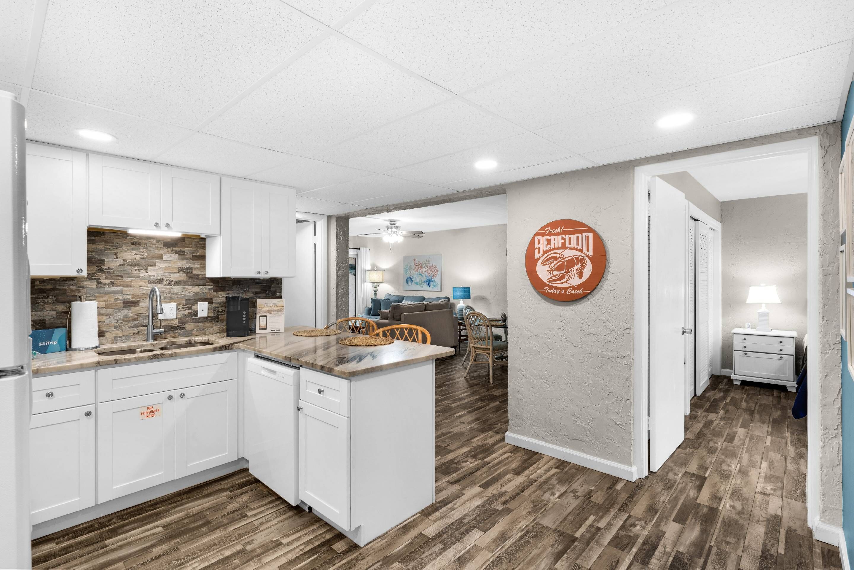 A modern kitchen with white cabinets, a stone backsplash, and a wooden floor. There is a dining area with a table and chairs, and a living room with a sofa and a coffee table.