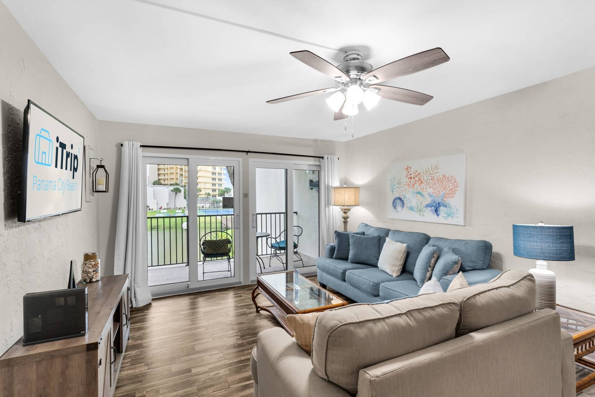 A living room with a blue sofa, a coffee table, a ceiling fan, and a view of a balcony with outdoor furniture.