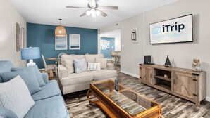 A modern living room with a blue accent wall, a ceiling fan, a large TV displaying 'iTrip Panama City Beach', and a wooden cabinet. - Spacious updated condo with a lake view, private balcony, and $300 Free Activities Daily! (Panama City Beach)