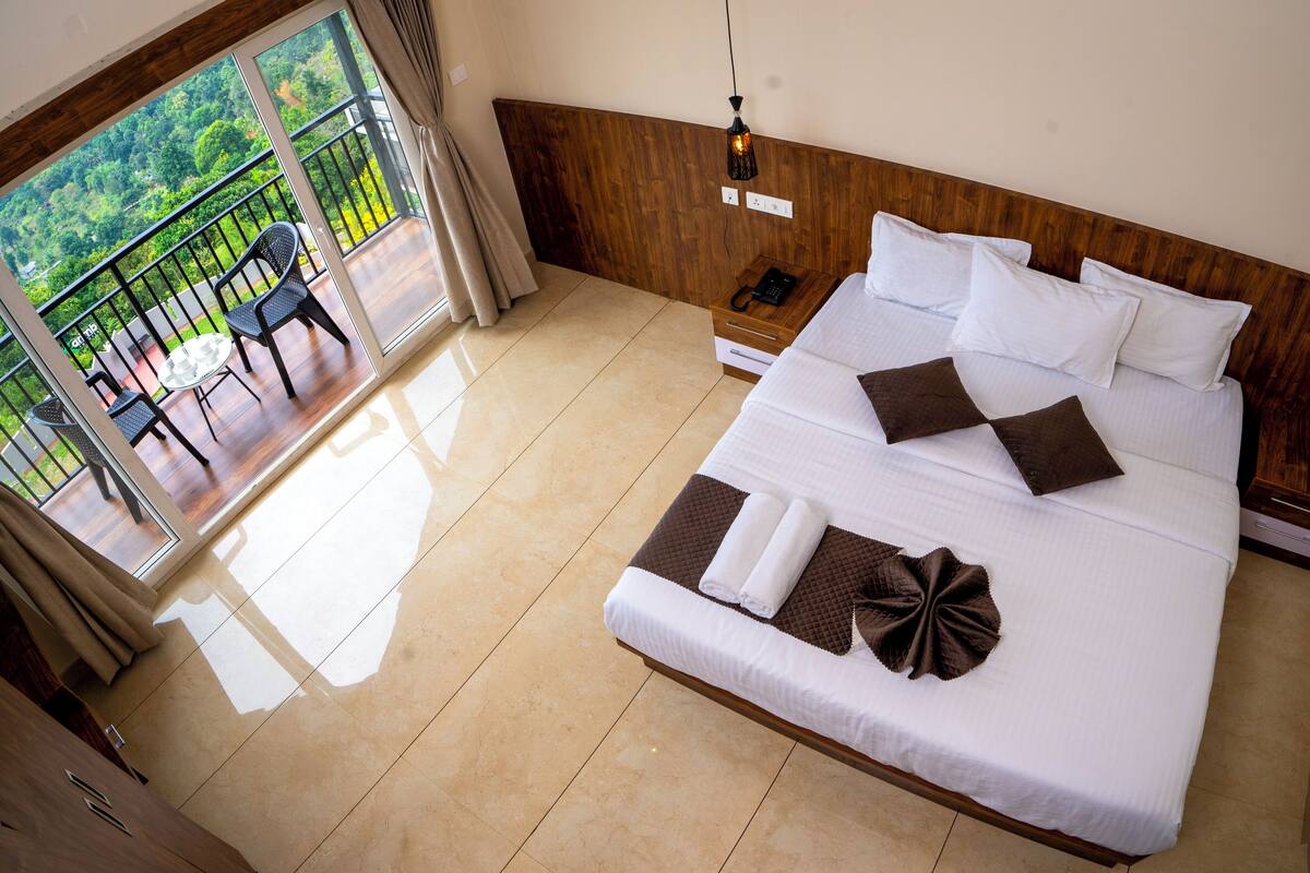 Une chambre d’hôtel avec un grand lit, une tête de lit en bois, un balcon avec des chaises et une vue sur la verdure.
