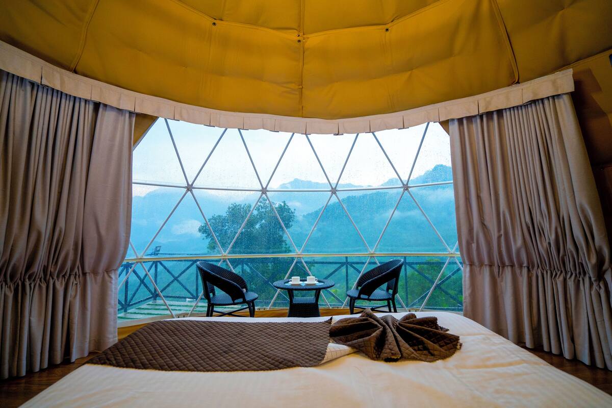 Une chambre avec un lit, deux chaises, une petite table et une vue sur les montagnes par une grande fenêtre.