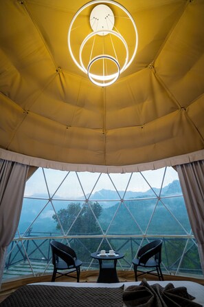 A tented room with a bed, two chairs, a table, and a large window offering a view of mountains. - Nexstay Karimb Resorts (Thamarassery)