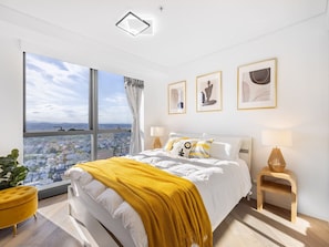 Une chambre moderne avec une grande fenêtre, un lit recouvert d’une literie blanche et d’un plaid jaune, et trois tableaux encadrés accrochés au mur.