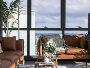 A modern living room with a brown leather sofa, a small round table with a vase and plant, and a large window with a view of the city.
