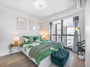 A modern bedroom with a large bed, a side table, a bench, and a view of the city through large windows.