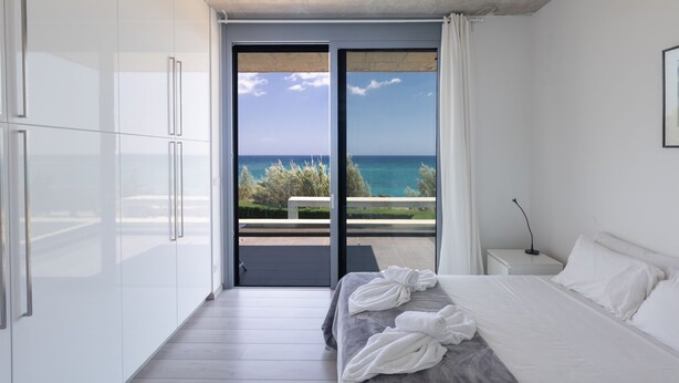 A modern bedroom with a large bed, white walls, and a view of the sea through a sliding glass door. - Sea front Elegant villa HEATED private POOL in Fig Tree Bay Protaras
  - 5 bedrm (Protaras)