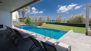 A modern outdoor pool area with a clear view of the ocean, surrounded by well-maintained greenery and a patio with lounge chairs. - Sea front Elegant villa HEATED private POOL in Fig Tree Bay Protaras
  - 5 bedrm (Protaras)