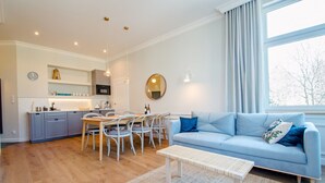 A modern living room with a blue sofa, a dining table with chairs, a kitchen area with cabinets and a sink, and a large window with curtains.