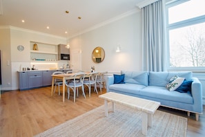 Ein modernes Wohnzimmer mit einem blauen Sofa, einem Esstisch mit Stühlen, einer Küchenzeile mit Schränken und einer Spüle sowie einem großen Fenster mit Vorhängen.